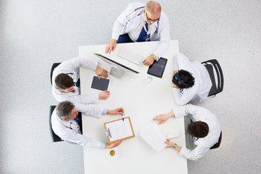 Medizinisches Team, bestehend aus 5 Ärzt:innen, sitzt und diskutiert am Tisch, man schaut aus der Vogelperspektive auf das diskutierende Ärzt:innenteam ©lenetstan/shutterstock.com